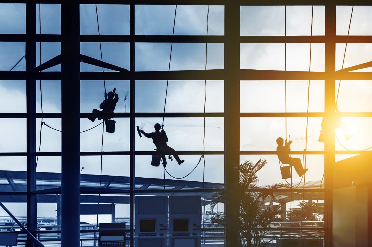 Window washers suspended on ropes clean large glass windows inside a sunlit airport terminal.